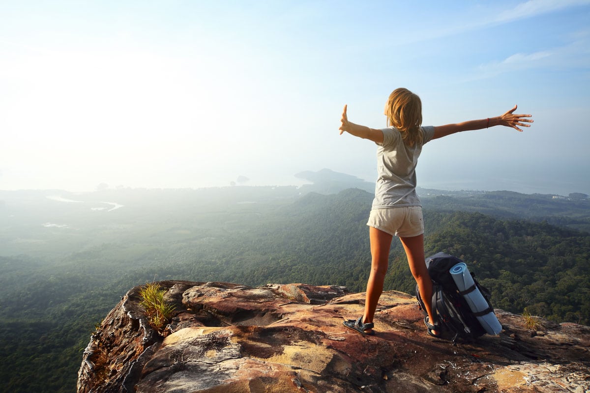 Young woman with backpack standing on cliff's edge and looking out on one of the vacations for over 50 singles
